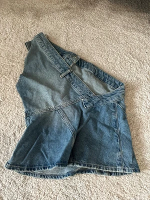 Blå jeans top från Zara, storlek S - Säljer en snygg jeans top från Zara i storlek S. Kjolen har en cool asymmetrisk design med flera sömmar och detaljer som ger den en unik look. Tillverkad i klassiskt blå denim med bälteshällor och knappstängning i midjan. 