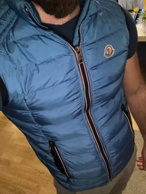 Blå dunväst Moncler, storlek S - Säljer en blå dunväst från Moncler med quiltad design och snygga detaljer vid dragkedjorna. Västen har Moncler-logga på bröstet och svarta insidor. Perfekt för dig som vill ha en stilren och sportig look.