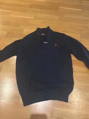Svart stickad tröja Polo Ralph Lauren. L - Snygg svart stickad tröja från Polo Ralph Lauren med röd broderad logga på bröstet. Tröjan har half zip och ribbad krage samt muddar. Perfekt för kyliga dagar och passar till jeans eller chinos.