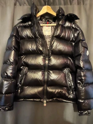 Svart Moncler pufferjacka med huva - Säljer en svart pufferjacka från Moncler med glansig finish och huva. Jackan har tvåvägsdragkedja med Moncler-logga, sidofickor med dragkedja och coolt serietryck på insidan. Perfekt för dig som vill ha en varm och snygg jacka med streetkänsla. Storlek 2 passar M 