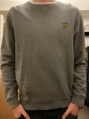 Grå sweatshirt från Lyle & Scott - Säljer en stilren grå sweatshirt från Lyle & Scott i storlek L. Tröjan har rund hals, lång ärm och den klassiska gula fågel-loggan på bröstet. Mjuk bomullskvalitet och ribbade muddar vid ärmslut och nederkant. Perfekt för chill dagar.