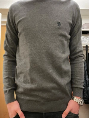 Mörkgrå tröja från U.S. Polo Assn. - Säljer en stilren mörkgrå tröja från U.S. Polo Assn. i storlek M. Tröjan har rund hals, diskret logga på bröstet och ribbade muddar vid ärmslut och nederkant. Materialet är mjukt och känns skönt mot huden, perfekt för dig som gillar klassisk och enkel stil.