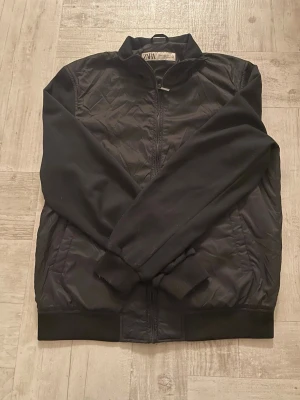 Svart bomberjacka från Zara - Säljer en svart bomberjacka från Zara i storlek M. Jackan har en klassisk rak passform, ribbade muddar och dragkedja framtill. Materialet är en mix av polyester och syntet, vilket gör den lätt och smidig. Perfekt för dig som gillar en clean och stilren look.
