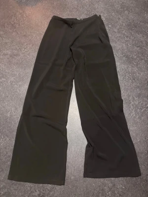 Svarta wide byxor i mjukt tyg - Säljer ett par svarta byxor med wide fit och rak siluett. Byxorna har hög midja och är gjorda i ett mjukt, flowy material som ger en skön känsla. Perfekta för dig som gillar en avslappnad och trendig look. Storlek 38, aldrig använda. Dragkedja i sidan.