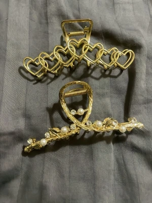 Guldiga hårklämmor med hjärtan och pärlor - Två snygga hårklämmor i guldfärgad metall. Ena klämman har ett mönster av sammankopplade hjärtan, medan den andra har dekorativa pärlor och små blommor längs kanten. Perfekt för att ge frisyren en extra touch av stil och elegans.