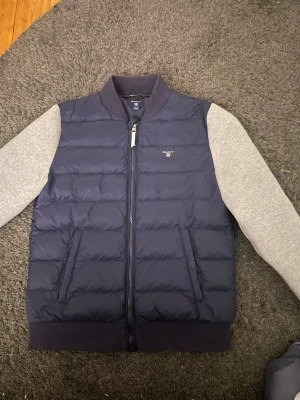 Mörkblå dunväst från GANT 170cm - Säljer en mörkblå dunväst från GANT med quiltad design och dragkedja framtill. Västen har en broderad GANT-logga på bröstet och ribbad krage. Insidan har texten 'GANT NEW HAVEN'. Perfekt för dig som gillar klassisk stil.