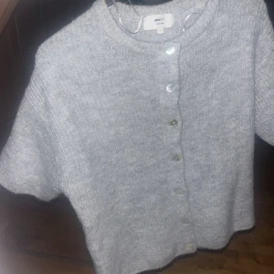 Grå stickad kofta från Gina Tricot - Mysig grå stickad kofta från Gina Tricot i storlek XS. Koftan har korta ärmar, rund hals och knäppning med fem pärlemorknappar framtill. Tillverkad i en mjuk ullblandning med ribbad struktur som ger en chill och modern vibe.