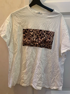 Vit t-shirt med leopardmönster Chiquelle - Säljer en vit t-shirt från Chiquelle i storlek M. T-shirten har ett stort leopardmönstrat tryck på bröstet och är klassiskt kortärmad med rund hals. Materialet är mjuk bomull och passformen är loose, perfekt för en chill och trendig look.