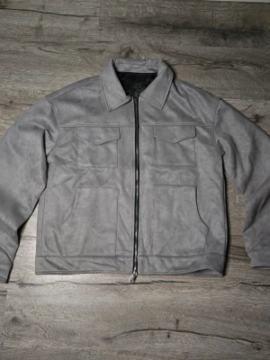 Grå bomberjacka - Snygg grå bomberjacka i mjuk polyester med två stora fickor framtill och dragkedja hela vägen. Jackan har klassisk krage och raka linjer, perfekt för dig som gillar stilren och enkel design. Fodret är gjort av terylene för extra komfort.