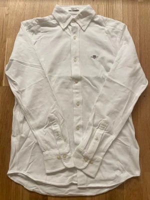 Skjorta från Gant  - Klassisk vit skjorta från Gant i Regular Fit och broderad logga på bröstet. Skjortan har långa ärmar, knappar framtill och är tillverkad i bomull för en skön känsla. Perfekt för fina tillfällen 