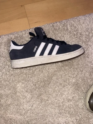 Adidas Campus sneakers marinblå - Säljer ett par Adidas Campus sneakers i marinblå mocka med klassiska vita ränder på sidorna och vit häl. Skorna har rund tå, platt sula och snörning framtill. Perfekt för dig som gillar retrostil och vill ha ett par snygga och bekväma sneakers. Endast använda 3 gånger , strolek 41,5. Tevka inte på att höra av dig om frågor eller funderingar!