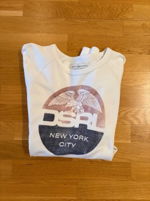 Vit långärmad tröja Ralph Lauren - Vit långärmad tröja från Denim & Supply Ralph Lauren med tryck i rött och blått framtill. Motivet har en örn och texten DSRL New York City. Rund hals och mjukt bomullsmaterial, perfekt för chill dagar.