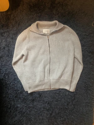 Grå stickad kofta från Maison Margiela - Säljer en grå heavyweight stickad kofta från Maison Margiela med ribbad struktur och hel dragkedja framtill. Koftan har en hög krage och långa ärmar, perfekt för lager på lager. Diskret Margiela-logga i nacken. Stilren och clean design som funkar till allt. Storlek s, lapp finns till tröjan