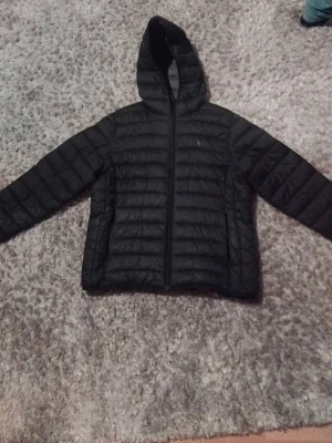 Svart pufferjacka med huva - Säljer en svart pufferjacka med huva och dragkedja framtill. Jackan har ett quiltat mönster och är långärmad, perfekt för kyliga dagar. Enkel och stilren design som passar till många olika outfits.
