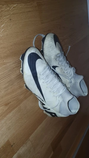 Nike Mercurial vita fotbollsskor - Säljer ett par vita Nike Mercurial fotbollsskor med svarta detaljer och stor Nike-logga på sidan. Skorna har snörning, högre strumpliknande skaft och är tillverkade i syntetmaterial. Perfekta för dig som vill sticka ut på planen.