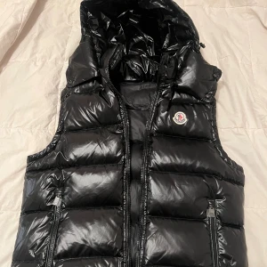 Svart dunväst från Moncler med huva - Säljer en svart, glansig dunväst från Moncler med huva och två dragkedjefickor. Västen har quiltad design och klassisk Moncler-logga på bröstet. Perfekt för dig som vill ha en snygg och varm look under höst och vinter. Dragkedja framtill och coola detaljer.
