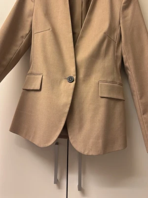 Beige kavaj från Zara - Snygg beige kavaj från Zara med klassisk passform och en knapp framtill. Jackan har två fickor med lock och är helfodrad. Perfekt för dig som vill ha en stilren look med enkel design och neutral färg. Den går in vid midjan och formar superfint! Färgen är finare i verkligenheten! 