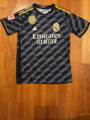 Real Madrid Adidas matchtröja 2024 - Säljer en officiell Real Madrid matchtröja från Adidas med FIFA World Champions 2024-märke. Tröjan har ett mörkblått och grått mönster, gula detaljer på axlarna och klubbens emblem på bröstet. Materialet är lätt polyester och designen är riktigt snygg för fotbollsfans.