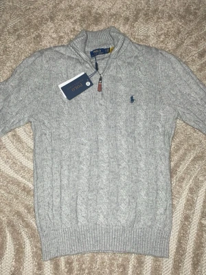 Grå stickad tröja från Polo Ralph Lauren - Snygg grå kabelstickad tröja från Polo Ralph Lauren med halv dragkedja och broderad marinblå logga på bröstet. Tröjan har långärmad design och ribbade muddar. Materialmix av ull, akryl och nylon för extra komfort och värme.
