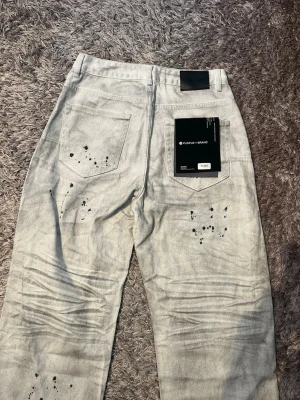 Ljusgrå jeans från Purple Brand - Säljer ett par ljusgrå jeans från Purple Brand med coola svarta färgstänk och skrynkliga detaljer på benen. Klassisk femficksmodell med silverfärgade nitar och knapp, samt en metallkedja vid hällan. Jeansen har en rak passform och är tillverkade i jeansmaterial.