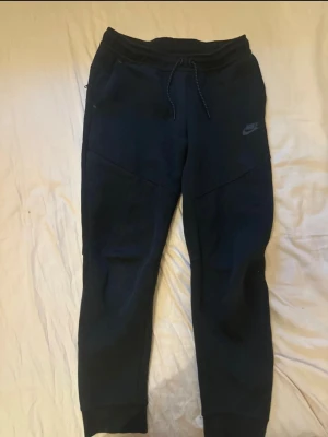 Svarta mjukisbyxor från Nike - Svarta mjukisbyxor från Nike med broderad logga på vänster ben. Byxorna har snörning i midjan, ribbade muddar vid benslut och praktiska fickor, varav en med dragkedja. Tillverkade i mjukt bomullsmaterial som är skönt att ha på sig. Perfekta för chill eller träning.