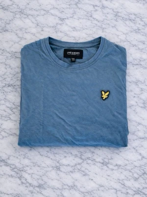 Blå t-shirt från Lyle & Scott - Uppgradera din vardagsstil med denna klassiska Lyle & Scott t-shirt i en elegant blå nyans. Tillverkad i mjuk, högkvalitativ bomull för en bekväm och lätt känsla hela dagen. Den diskreta, ikoniska gyllene örnlogotypen på bröstet ger en subtil men stilren detalj som gör tröjan både tidlös och trendig. Perfekt att bära ensam eller under en jacka för en avslappnad, men ändå sofistikerad look. Färg: Blå Material: 100% bomull Passform: Regular fit Detaljer: Broderad gyllene örnlogotyp på bröstet 