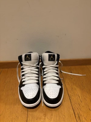 Nike Air Jordan 1 Mid svart/vit/guld - Säljer ett par Nike Air Jordan 1 Mid sneakers i svart, vit och guld. Skorna har klassisk snörning, perforerad tå och guldiga detaljer på swoosh och logga. Tillverkade i skinn med platt sula och rund tå. Perfekt för dig som vill ha en ikonisk och stilren sneaker.