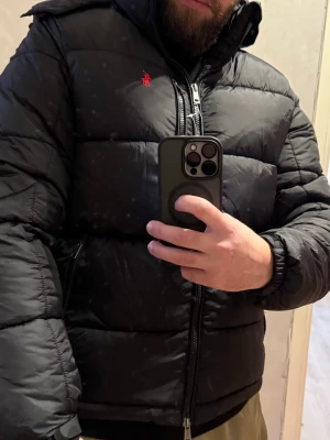 Svart pufferjacka från Polo Ralph Lauren - Svart pufferjacka från Polo Ralph Lauren med röd broderad logga på bröstet. Jackan har huva, dragkedja framtill och elastiska muddar vid ärmslut. Materialet är syntet och jackan är quiltad för extra värme. Perfekt för kalla dagar.