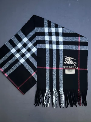 Rutig svart halsduk från Burberry - Snygg svart halsduk från Burberry med klassiskt rutigt mönster i grått, vitt och röda linjer. Halsduken har fransar längst ner och en broderad Burberry-logga med riddare på framsidan. Materialet är mjuk kashmir aktigt som känns lyxigt och skönt mot huden.