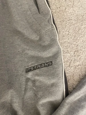 Ietsfrans - Säljer ett par grå mjukisbyxor från Itsfrans med svarta sidopaneler som ramas av vita kanter! Dom är helt nya, lappen är borta och använda endast 1 gång. Byxorna är gjord av bomull och midjan väldigt strechig. Två stora rymliga fickor framtill, och en baktill.💕