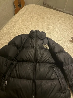 Svart pufferjacka från The North Face - Säljer en svart pufferjacka från The North Face i storlek M. Jackan har hög krage, två sidofickor med dragkedja och tydligt logotryck på bröstet och ryggen. Materialet är syntet och dun, perfekt för kalla dagar. Stängs med dragkedja framtill. Org pris är 3945kr, köpt förra året knappt använt. 