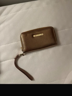 Guld plånbok från Style med dragkedja - Snygg guldplånbok från Style med praktisk handledsrem och dragkedja runtom. Plånboken har flera fack för kort och sedlar, perfekt för att hålla ordning på allt viktigt. Stilren design med metallmärke framtill och mjukt skinnliknande material.