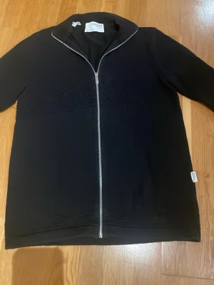 Svart kofta från Selected Homme - Svart kofta med hel dragkedja från Selected Homme. Tröjan har ett strukturerat stickat mönster över bröstet och axlarna, samt en hög krage. Materialet känns mjukt och har en modern look, perfekt för dig som gillar stilrena plagg med lite edge.