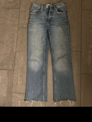 Blå croppade jeans från Zara - Säljer ett par blå jeans från Zara med croppad passform och råa, fransiga benslut. Jeansen har klassisk femficksdesign, hög midja och dragkedja med knapp. Materialet är jeans i bomull med lätt tvättad look och ljusare partier på benen. Byxan sitter lite löst under kedjan