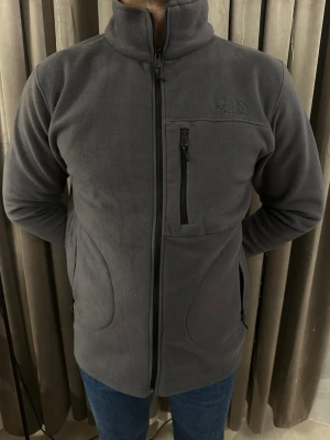 Grå fleecejacka från The North Face - Mjuk och varm grå fleecejacka från The North Face med hög krage och hel dragkedja framtill. Jackan har tre fickor med dragkedja, varav en på bröstet, och diskret logga på vänster sida. Perfekt för kyliga dagar och enkel att matcha med jeans.