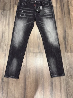 Svarta Dsquared2 jeans med slitningar - Säljer ett par svarta jeans från Dsquared2 med coola slitningar och blekta detaljer. Modellen har raka ben, klassisk femficksdesign och Dsquared2-logga bak i midjan. Jeansen är gjorda i jeansmaterial och har en snygg streetstil.