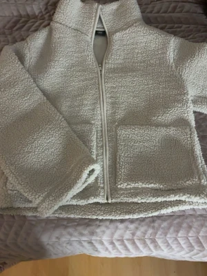 Beige teddyjacka från lager 157, storlek S - Mysig beige teddyjacka från lager 157 i storlek S. Jackan har dragkedja framtill, två stora fickor och hög krage. Perfekt för kyliga dagar när du vill vara både varm och stilren.
