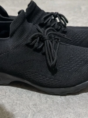 Svarta stickade sneakers strl 37 - Svarta sneakers med stickad ovandel och snörning. Skorna har en rund tå och platt sula, vilket gör dem riktigt sköna att ha på sig. Materialet är syntet och de har en stilren, helsvart look som passar till många outfits.