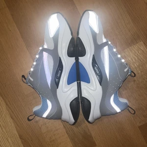 Dior b22 - Bara seriösa köpare tack!. Säljer ett par oanvända Dior b22s med cool design i grått, vitt och blått. Skorna har reflekterande detaljer, meshpartier och vita snören. Sulan är tjock och svart med vita inslag, och insidan har tydlig Dior-logga. Perfekt för dig som gillar exklusiva och trendiga sneakers. 