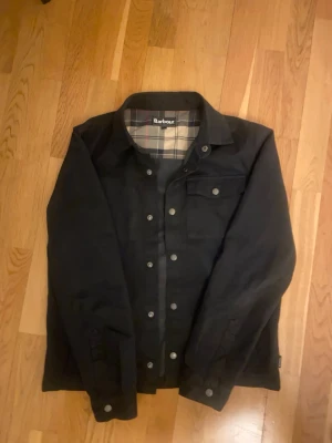 Svart jacka från Barbour - Svart jacka från Barbour i storlek S. Klassisk modell med knappar framtill och en bröstficka med lock. Insidan har ett snyggt rutigt foder i beige, blått och rött. Perfekt för dig som gillar stilrena och tidlösa plagg.