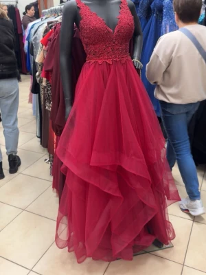 Röd långklänning med spets och volang - Säljer en fantastisk röd långklänning med v-ringad spetsliv och volangig kjol i flera lager. Klänningen har smala axelband och en elegant, luftig känsla. Perfekt för bal eller festliga tillfällen. Materialet är lätt och ger ett riktigt lyxigt intryck.                                                                        Helt ny oanvänd balklänning, köpt som reserv men bestämt mig för en annan.