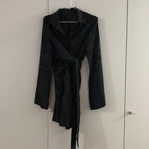 Svart omlottklänning med blommönster - Snygg svart kortklänning med subtilt blommönster och omlottdetalj i midjan. Klänningen har långärmade ärmar, krage och knappar framtill. Materialet har en lätt glansig finish som ger en exklusiv känsla. Perfekt för dig som gillar stilrena och trendiga plagg. Går att justera storleken med bandet 