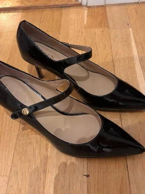 Svarta klackskor från Lauren Ralph Lauren - Snygga svarta klackskor i lack från Lauren Ralph Lauren med spetsig tå och smal klack. Skorna har en elegant rem över foten med guldfärgad knappdetalj. Perfekt för dig som gillar klassisk och stilren design. De har en skada på ena klacken. Orginalpris 1800 kr. 