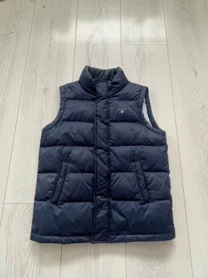 Mörkblå dunväst från GANT - Säljer en mörkblå dunväst från GANT med quiltad design och ståkrage. Västen har två fickor framtill och dragkedja. Insidan är fodrad med blåvita ränder för en snygg design! Vid intresse skicka! 