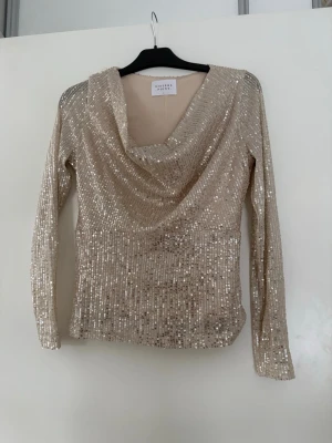 Glittrig långärmad topp Sisters Point - Säljer en glittrig champagnefärgad topp från Sisters Point i storlek S. Toppen har långa ärmar, paljetter över hela ytan och ett snyggt draperat fall framtill. Materialet är polyester med elastan för stretch och komfort. Perfekt för att sticka ut med extra bling. Endast testad!! Kan saknas lite paljetter på något ställe 