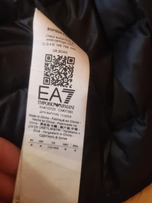 Svart pufferjacka EA7 Emporio Armani - Svart pufferjacka från EA7 Emporio Armani med huva och guldfärgat logotryck på bröstet. Jackan har quiltad design, dragkedja framtill och långa ärmar. Perfekt för dig som vill ha en snygg och stilren look under kyliga dagar. Pris kan diskuteras då jag vill bli av med den