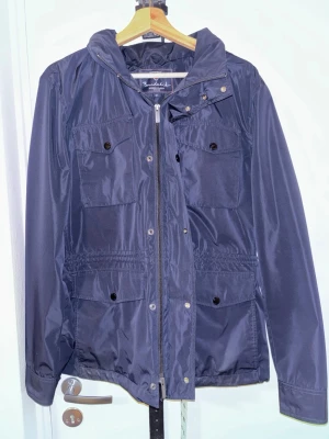 Mörkblå fältjacka från Bondelid - Snygg mörkblå fältjacka från Bondelid med fyra framfickor med knappar och dragkedja framtill. Jackan har huva och är tillverkad i glansig polyester som ger en stilig vibe. Perfekt för dig som gillar klassisk stil med modern touch.