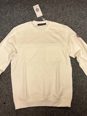 Vit sweatshirt från Canada Goose - Säljer en stilren vit sweatshirt från Canada Goose med rund hals och en praktisk bröstficka. Tröjan har långärmade ärmar och en diskret logotyp på vänster ärm. Perfekt för dig som gillar enkel och clean design.
