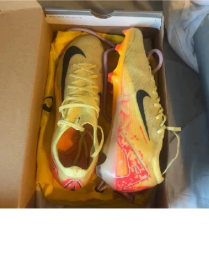 Nike Mercurial Air Zoom fotbollsskor - Säljer ett par Nike Mercurial Air Zoom fotbollsskor i ljusgul med svarta och röda detaljer. Skorna har snörning, platt sula med dobbar och är tillverkade i syntetmaterial för grym komfort och grepp på planen. Snygg design med pixelmönster och medföljande gul skopåse.