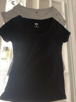 Svart och grå t-shirt från 157 XS - Två basic t-shirts från 157 i storlek XS, en i svart och en i grått. Båda har rund hals och korta ärmar, perfekta för att bygga lager eller köra solo. Materialet är mjukt med bomull och lite elastan för skön stretch. Snygga och enkla att matcha med allt.